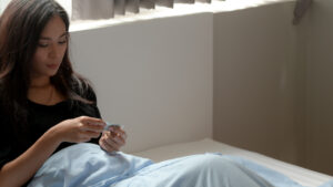 Photo of a young woman sitting in a bed with sheets over her legs, considering whether or not to take a Misoprostal pill.